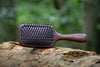 Root Ritual sandalwood paddle scalp brush resting on a forest log