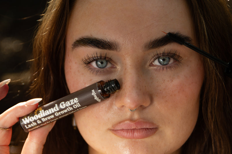 Woman holding Woodland Gaze lash and brow growth oil while brushing it through her brows